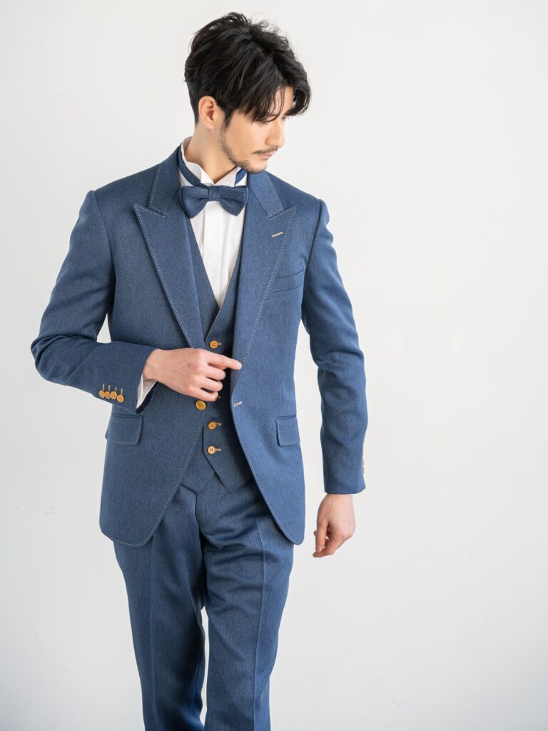 AMM-021(ネイビー)| 「ナチュレミア」という新素材を使ったデニムブルーのタキシード。デニムに見えますがデニムとは違い、 ポリエステル100%でありながら、天然素材のような肌触りと質感を持ち吸水性と速乾性、シワになりにくい強度を兼ね備えた大変扱いやすい生地で仕立てています。 手入れがしやすく、永く使うことができる世界的なSDG'sの潮流を汲んだ新素材です。 独特の色目で、カジュアルさの中にこだわりとおしゃれ要素が内包された ハイブリッドなデザインがご好評をいただいております。 また、ベージュのリバーシブルベストで印象を変えて、気軽におしゃれを楽しむようにお召しいただけます。