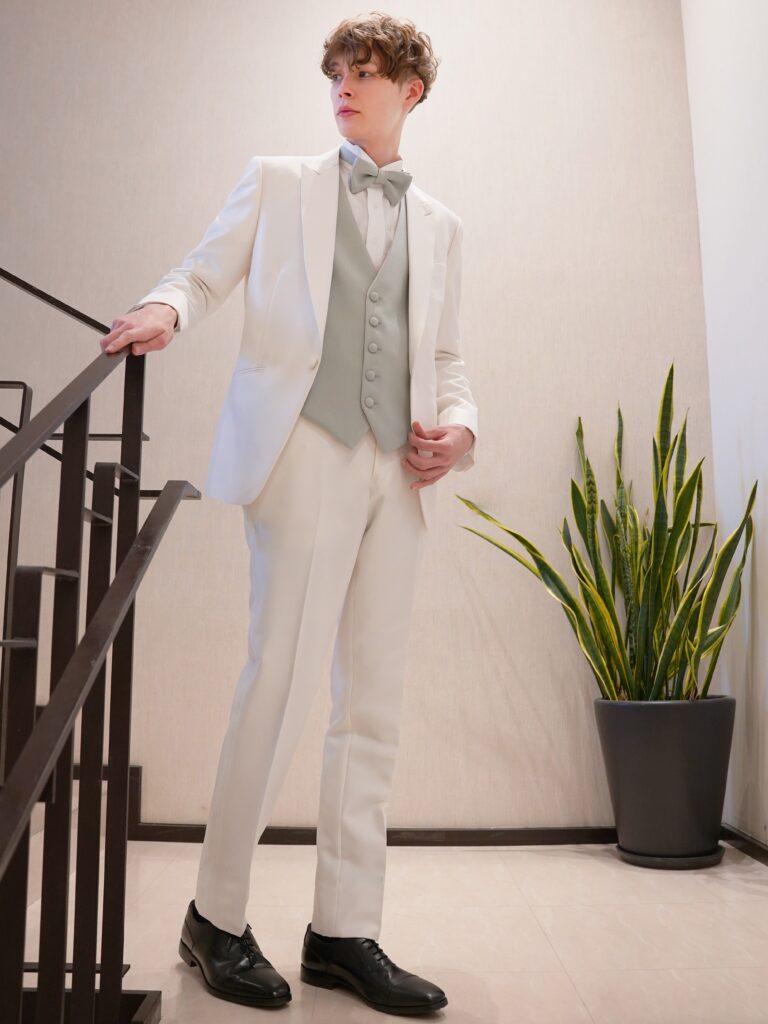 AMM-029(ホワイト) | 艶感が際立つピュアホワイトのタキシードが、圧倒的な存在感と上質さを放つ一着。 繊細な光沢をたたえる生地は、フォーマルでありながら華やかさも兼ね備え、特別な一日にふさわしい仕上がりです。 インナーには、グリーンとベージュのリバーシブルベストをコーディネート。 洗練されたグリーンは落ち着いた気品を演出し、ベージュにチェンジすれば柔らかで上品な印象へと表情を変えます。 一着でふたつの魅力を持つこのスタイリングは、昼夜での使い分けやシーンチェンジにも最適。 クラシックとモダンが調和する、こだわりを感じさせるタキシードスタイルです。