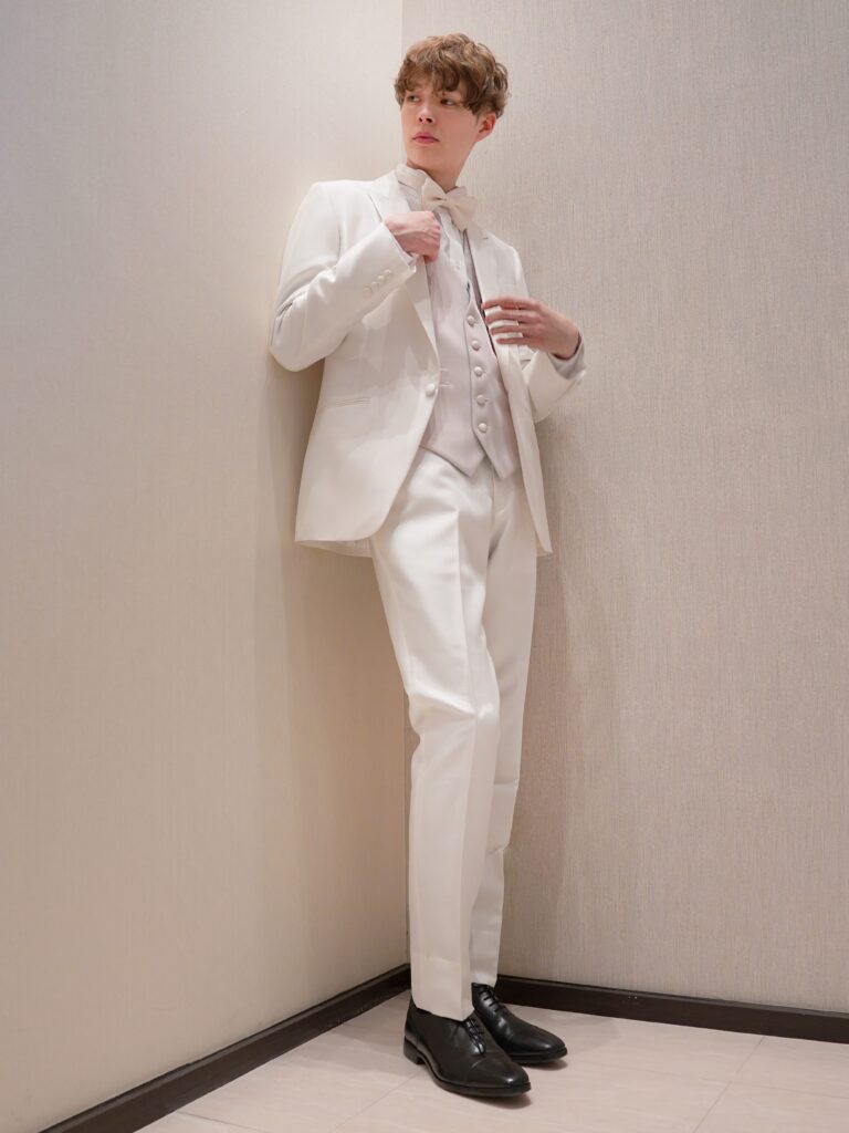 AMM-029(ホワイト) | 艶感が際立つピュアホワイトのタキシードが、圧倒的な存在感と上質さを放つ一着。 繊細な光沢をたたえる生地は、フォーマルでありながら華やかさも兼ね備え、特別な一日にふさわしい仕上がりです。 インナーには、グリーンとベージュのリバーシブルベストをコーディネート。 洗練されたグリーンは落ち着いた気品を演出し、ベージュにチェンジすれば柔らかで上品な印象へと表情を変えます。 一着でふたつの魅力を持つこのスタイリングは、昼夜での使い分けやシーンチェンジにも最適。 クラシックとモダンが調和する、こだわりを感じさせるタキシードスタイルです。