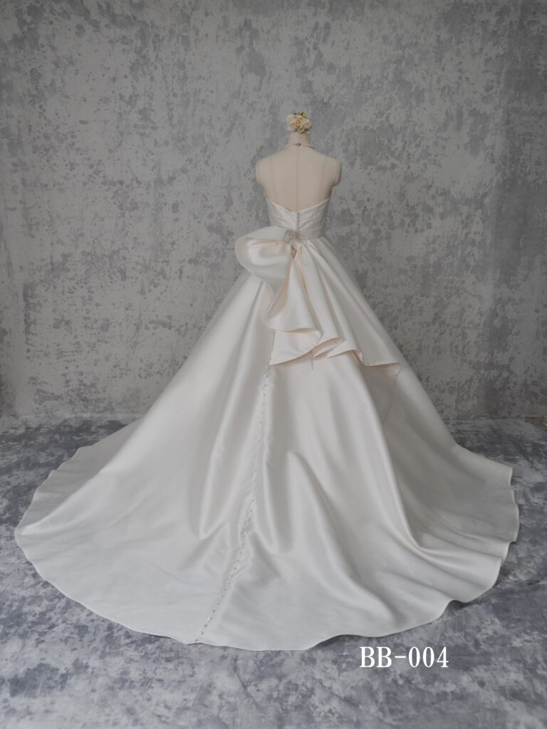 BB-004 | 上品な光沢を放つミカド素材を全面に用いたシンプルなマリエ。幾重にも重ねられてボリューム感を表現したスカートシルエット、ウェストラインにはアシメトリーにタックを取ったサッシュベルト、足長効果を生みます。バックにはアシメトリーの大きなリボンが、スカート裾まで続くクルミボタンと相俟って、シンプルな中に印象的なバックデザインに。