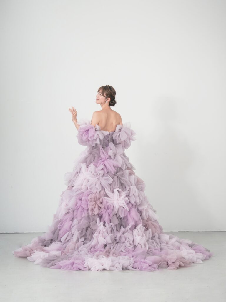 LQ-027(ラベンダー) | 柔らかいピンク・ラベンダーの色を複数混ぜ合わせてひとつずつふんわりと、立体的に形成したハンドメイドフラワーをそのまま身に纏うようなフォトジェニックなドレス。花びらを一枚一枚つまんでふんわり見せる工程や、お花中心のクリスタルをチュールでくるむ技法などよく見るととっても繊細な手仕事でできているのがわかります。