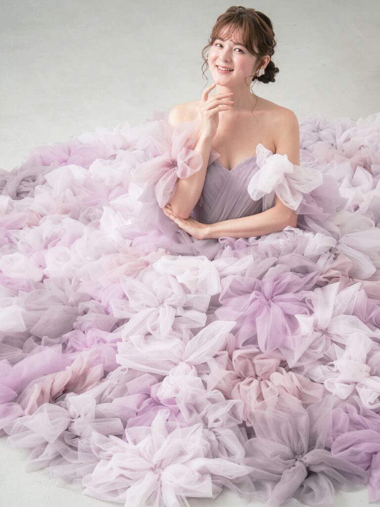 LQ-027(ラベンダー) | 柔らかいピンク・ラベンダーの色を複数混ぜ合わせてひとつずつふんわりと、立体的に形成したハンドメイドフラワーをそのまま身に纏うようなフォトジェニックなドレス。花びらを一枚一枚つまんでふんわり見せる工程や、お花中心のクリスタルをチュールでくるむ技法などよく見るととっても繊細な手仕事でできているのがわかります。