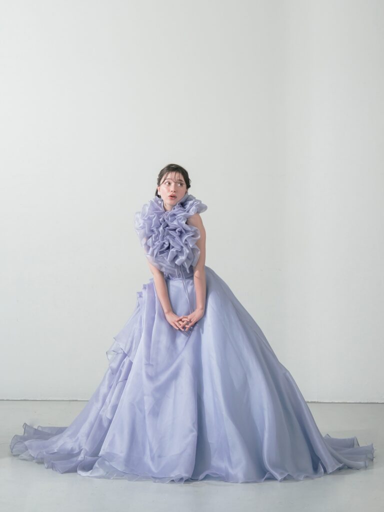 ST-111 | やわらかなウィスティリアカラーが空気まで優しく染める一着。透明感のあるオーガンジーの重なりと、流れるようなボリュームシルエットが、動くたびに表情を変えてくれます。 甘すぎず、上品で、記憶に残るカラードレス。静かな華やかさをまといたい花嫁さまへ。